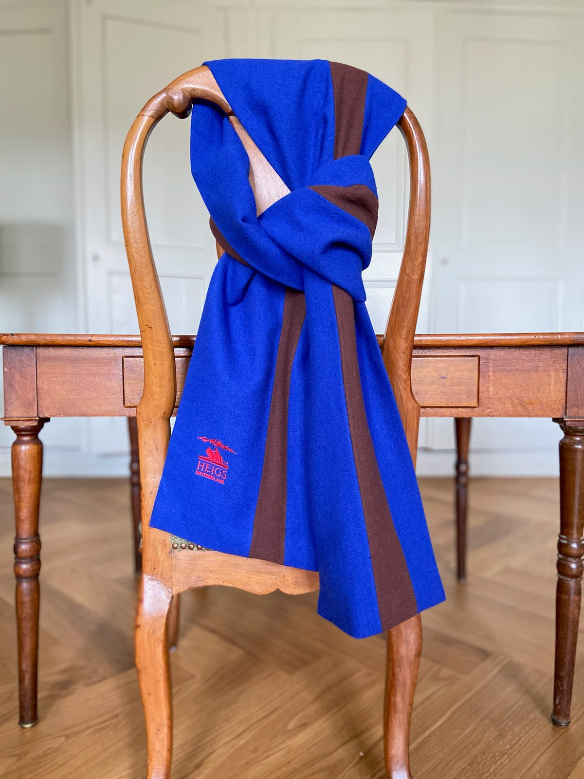 The University Scarf in royal blue and tobacco wool, knotted over a wooden chair — red HEIGS embroidery at the hem