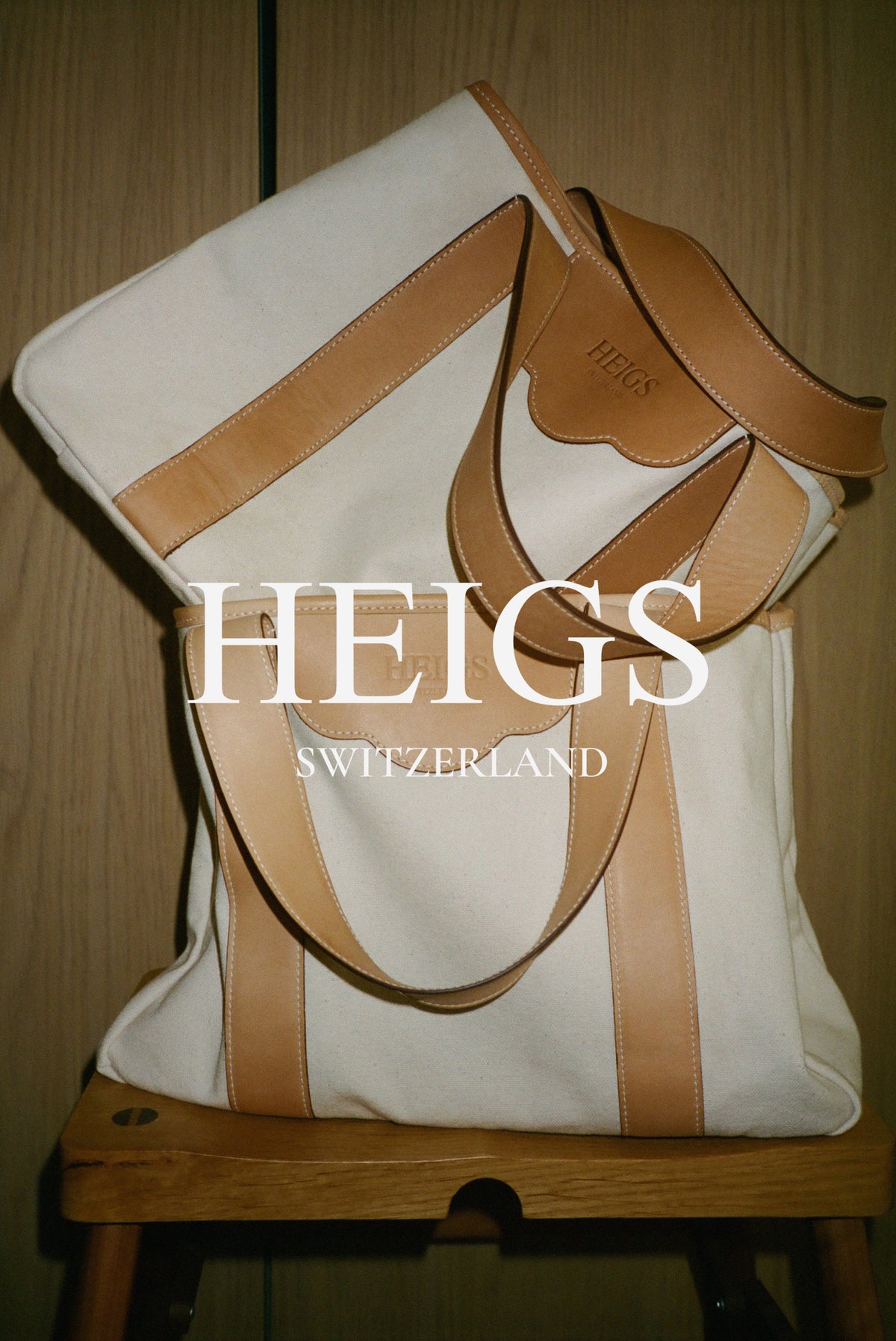 Two HEIGS à New York tote bags in Natural, cream canvas with tan leather handles, stacked on a wooden stool